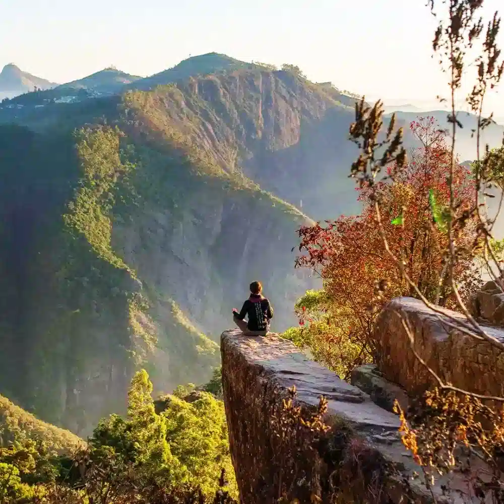A person meditating on the edge of Dolphin's Nose cliff in Kodaikanal, overlooking deep valleys and misty mountains.