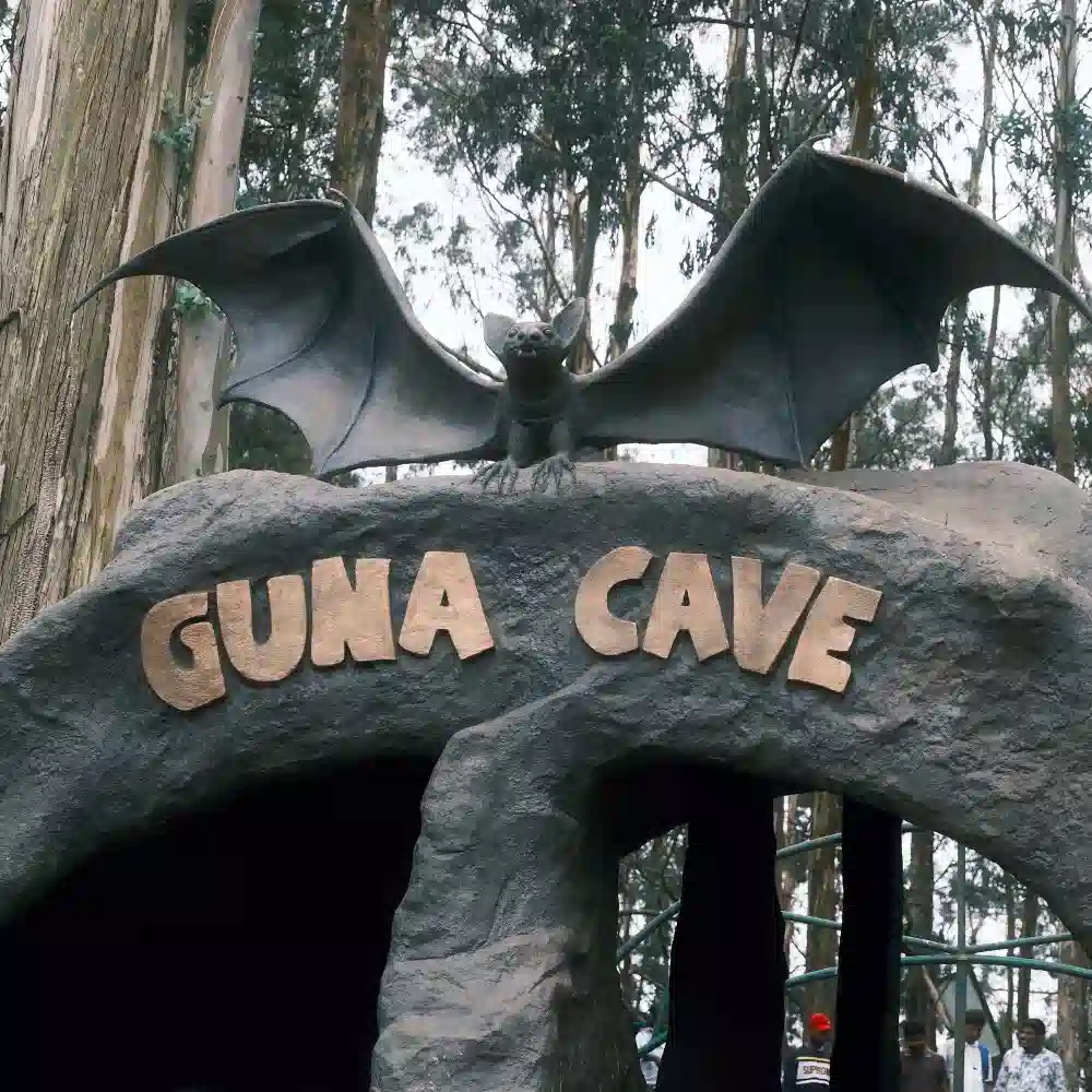The entrance sign for Guna Caves in Kodaikanal featuring a large bat sculpture on top of a rock archway in the woods.