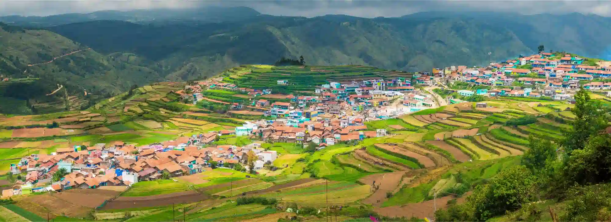 A wide panoramic view of Poombarai village in Kodaikanal showing colorful houses nestled among green terrace farm fields.