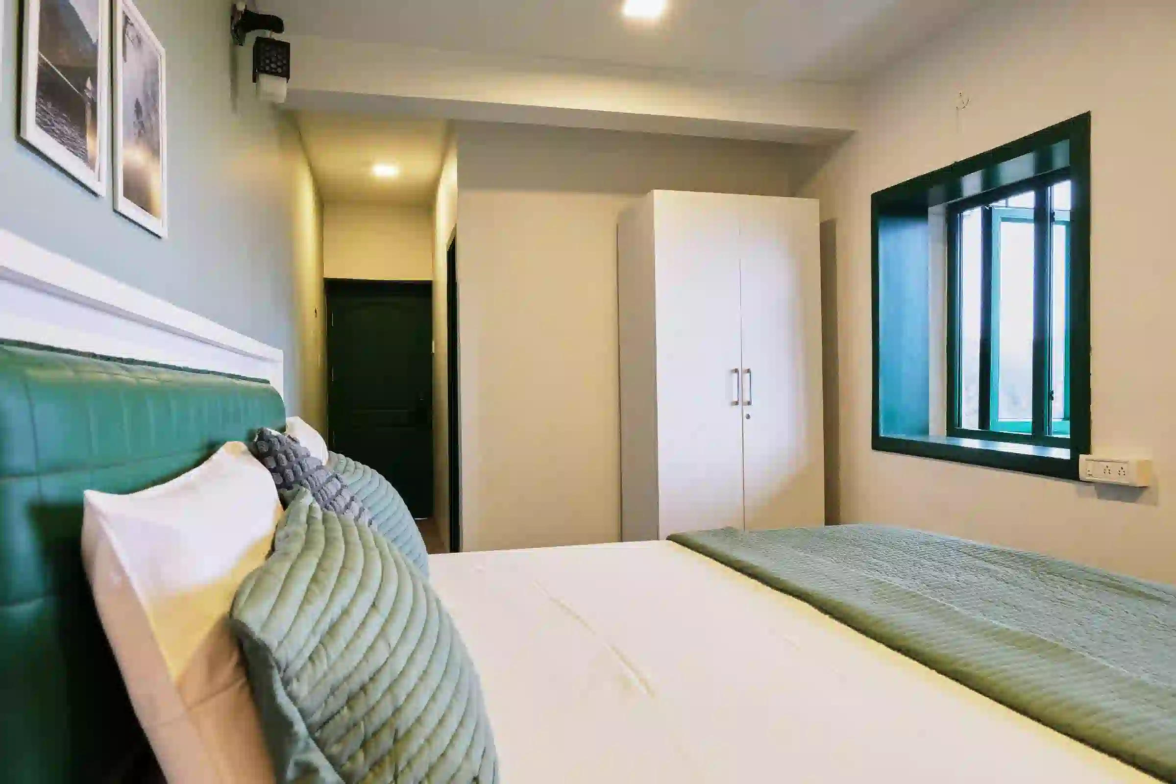 Cardamom Pods couple room featuring a white bed with green headboard, large wardrobe and window with dark green frames.